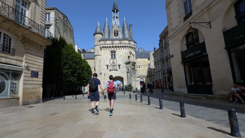 Andrew Curry and Dylan Cunningham holding hands walk toward the Porte Cailhau, a medieval gate with pointed turrets, in Bordeaux, France