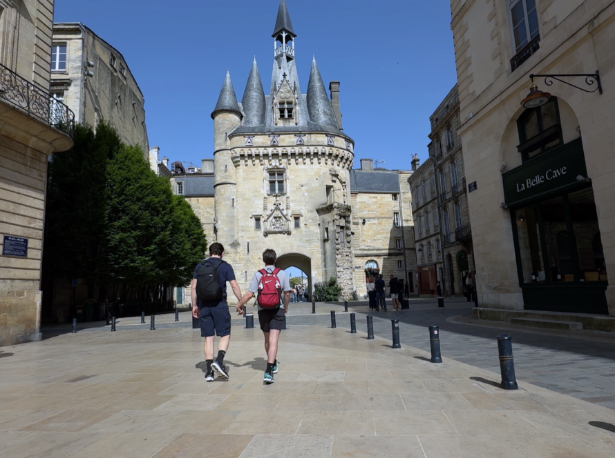 Andrew Curry and Dylan Cunningham holding hands walk toward the Porte Cailhau, a medieval gate with pointed turrets, in Bordeaux, France