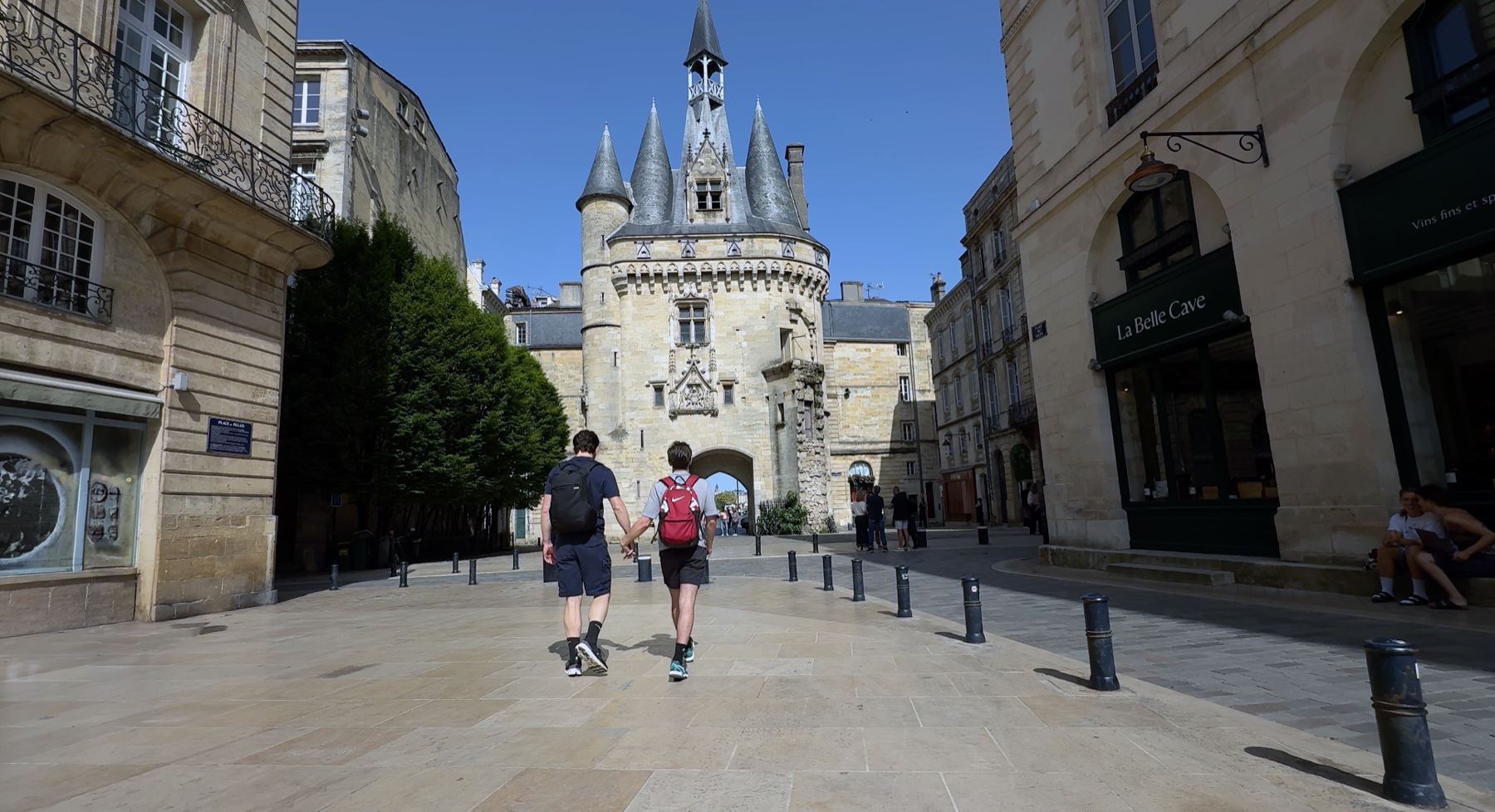 Andrew Curry and Dylan Cunningham holding hands walk toward the Porte Cailhau, a medieval gate with pointed turrets, in Bordeaux, France